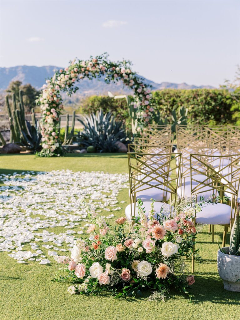 Outdoor wedding ceremony space with arch of greenery and white, blush, pink, and mauve flowers, aisle rose petals and back of ceremony ground arrangements.