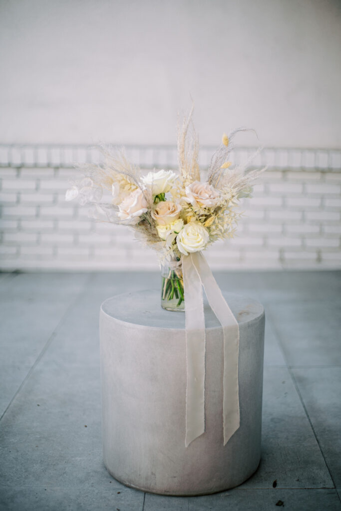 Bridal bouquet in glass vase on cement pillar of white and soft blush roses with pampas, dried bunny tales, and dried lunaria.