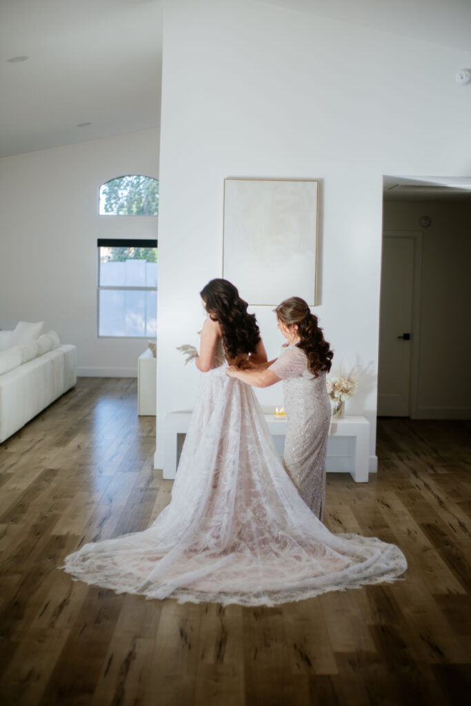 Bride getting ready into dress with another woman's help buttoning up back in large room of a home.