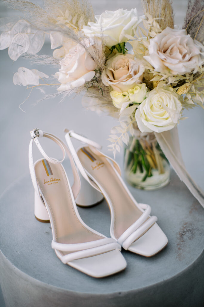 White bridal shoes on cement pillar next to bouquet of neutral floral and pampas in glass vase.