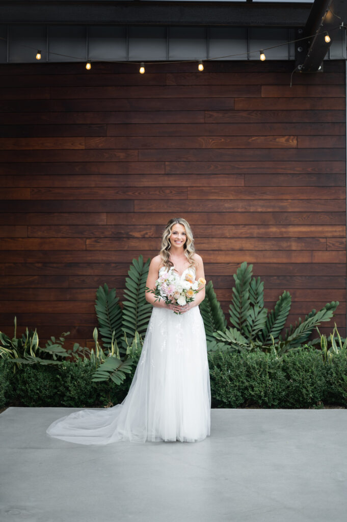 Bride standing in front of dark wood wall with green plants on ground, smiling, holding bouquet.