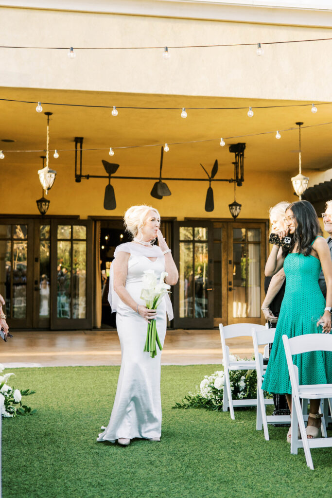 Bride blowing a kiss while walking down wedding ceremony aisle holding bouquet of calla lilies.