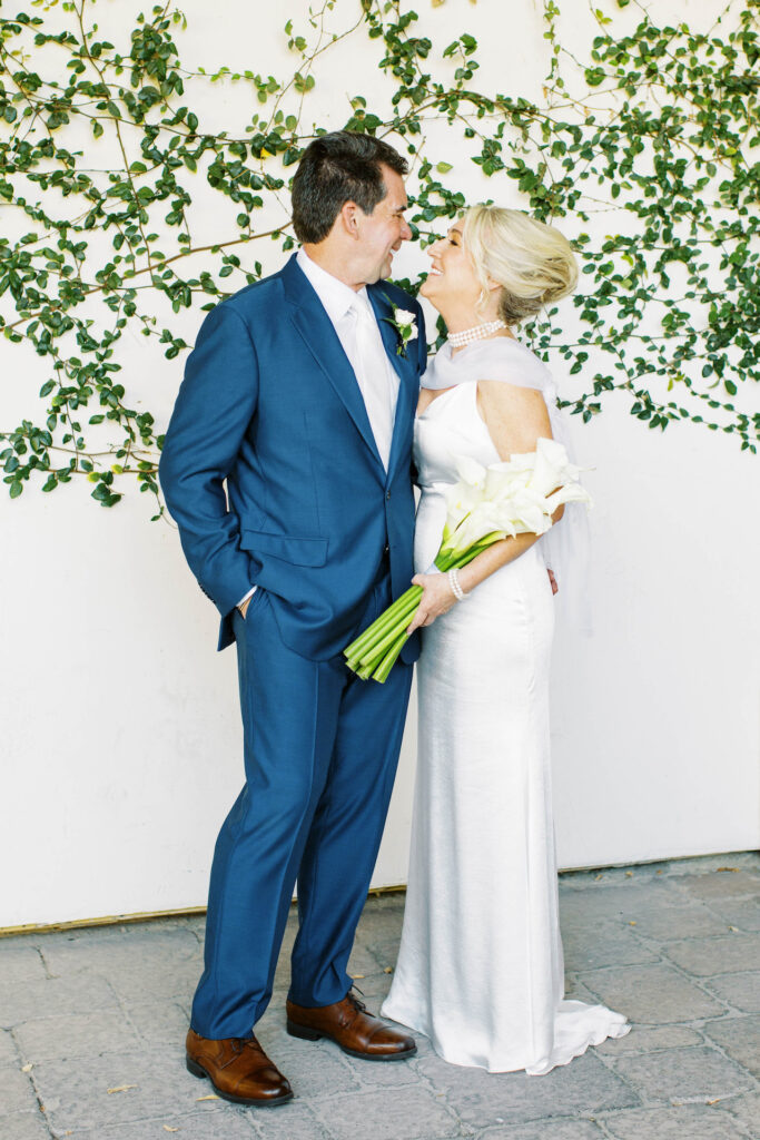 Bride and groom smiling at each other in front of white wall with vines growing on it, bride holding bouquet of calla lilies.