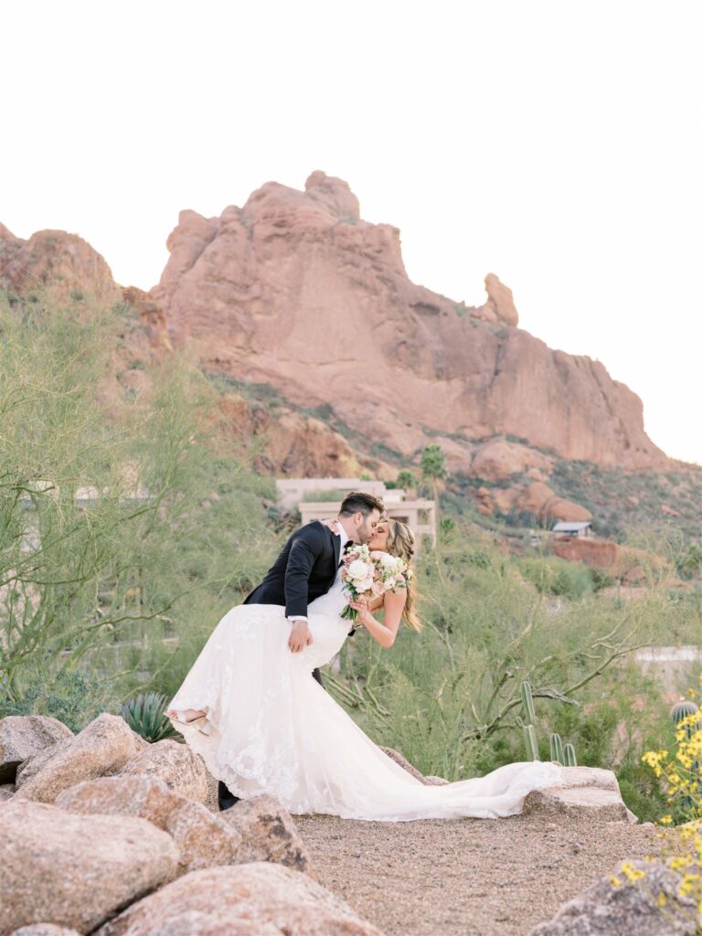 Groom dipping bride while they are kissing each other in desert landscape with mountain in the background.
