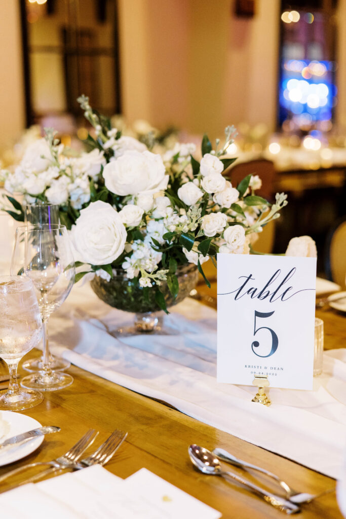 Large wedding reception centerpiece of white flowers and greenery in a glass vase next to table number.