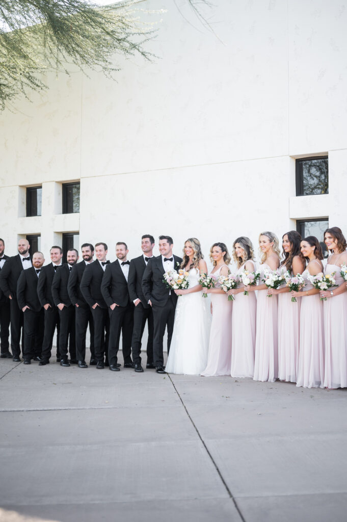 Wedding party standing in a line, smiling. Men in black with bowties and women in soft pink dresses.