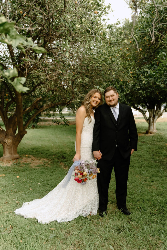 Bride and groom standing in grass yard with trees, holding hands, smiling.