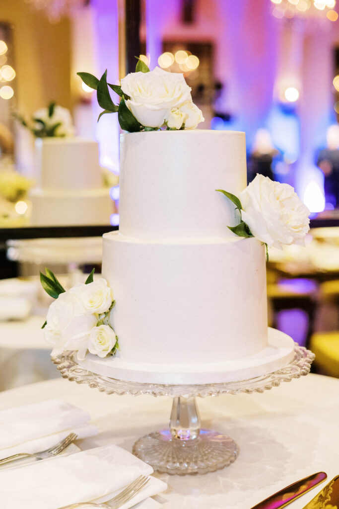 Two tiered white wedding cake with white roses added.