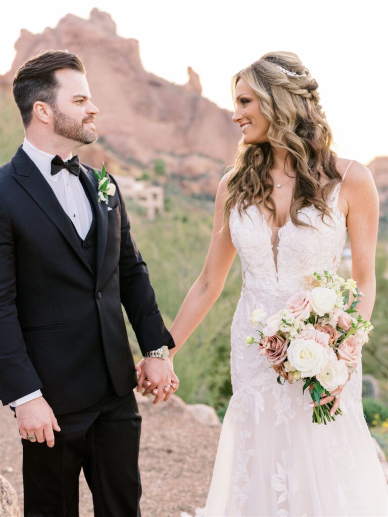 Bride and groom holding hands, smiling at each other while standing in desert landscape.