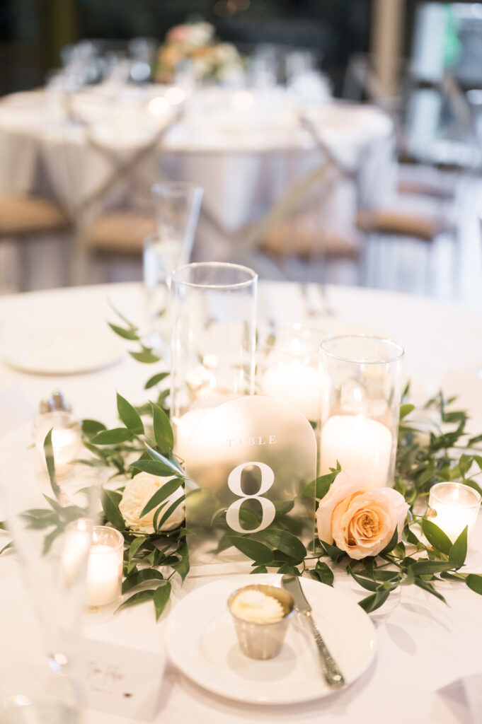 Reception table centerpiece of frosted table number sign with greenery and light pink roses and pillar candles.