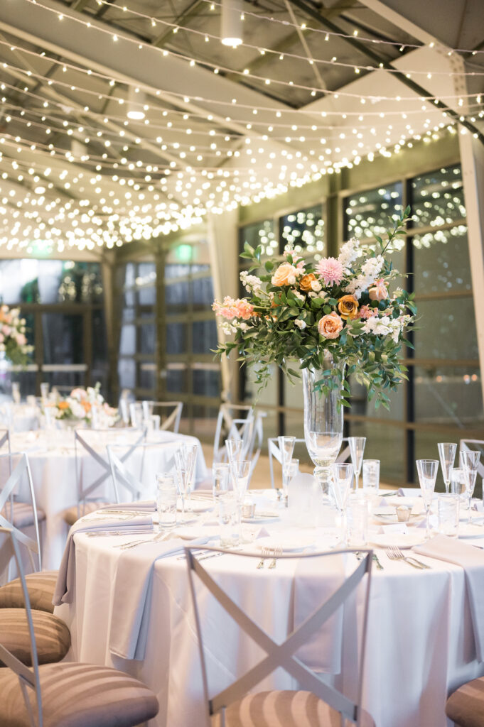 Round reception tables at indoor event with twinkle lights strung across ceiling. Tall and low floral centerpeices.