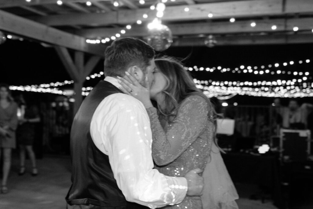 Bride and groom kissing under twinkle lights at wedding reception on dance floor.