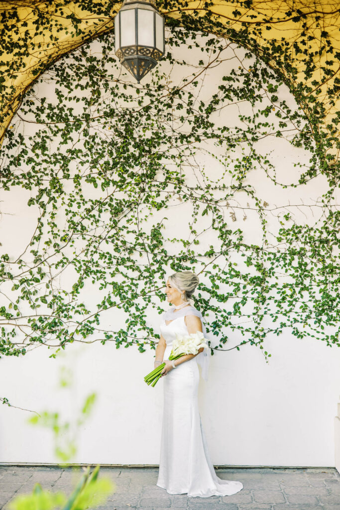 Bride standing in front of white wall with vines growing on it, holding bouquet of calla lilies.
