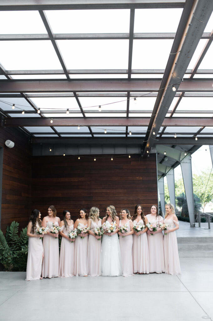 Bridesmaids in blush pink dresses standing in a line with bride, smiling and looking at each other.