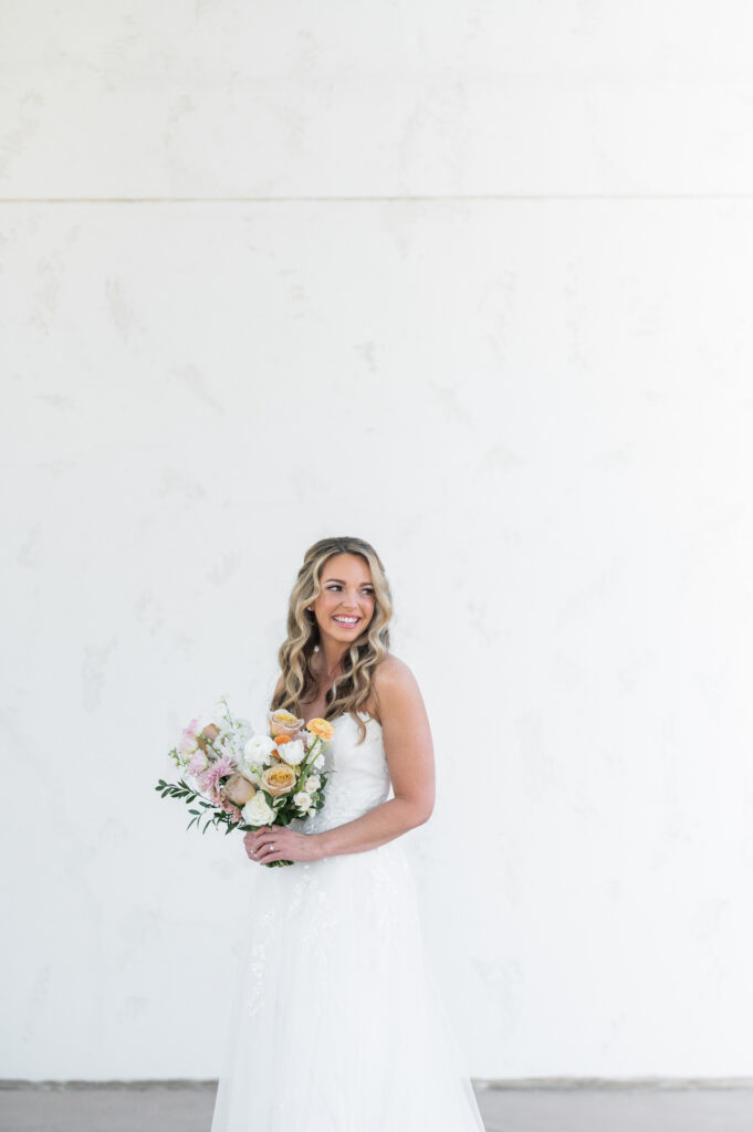 Bride looking over shoulder smiling while holding bouquet standing in front of white wall.