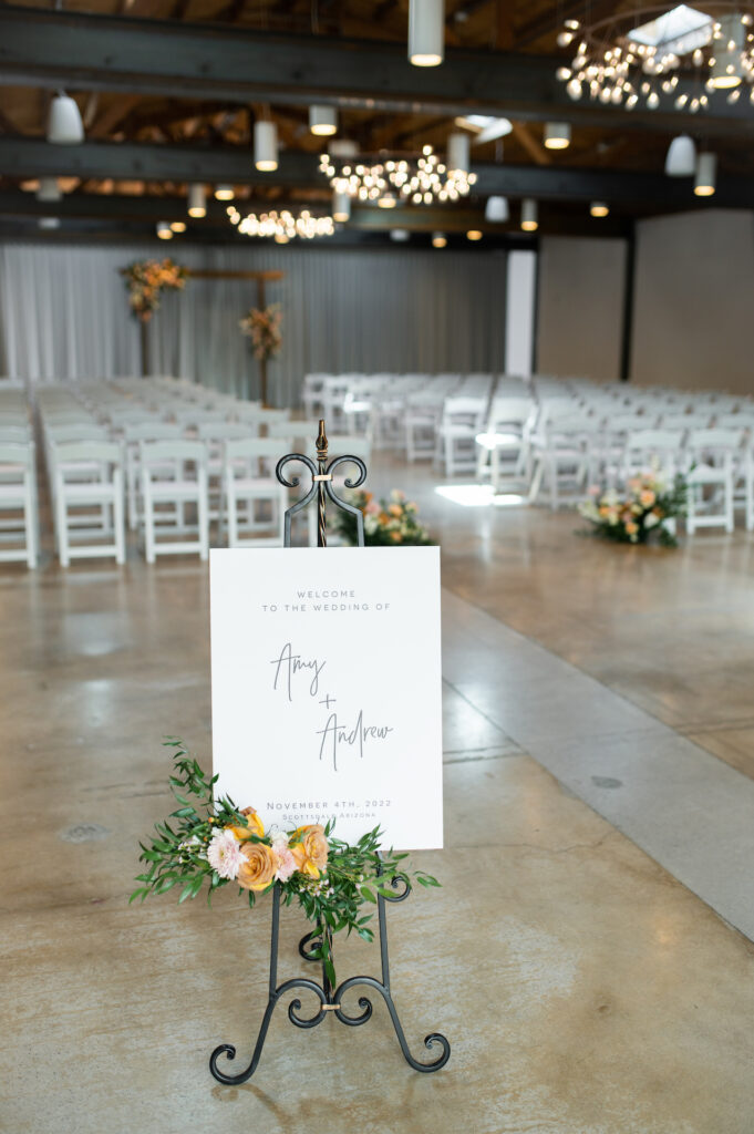 White welcome sign to indoor wedding ceremony on easel with floral spray at bottom of sign.