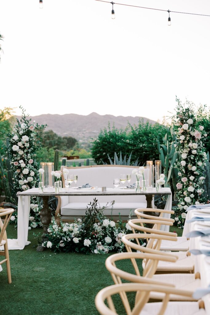 Sweetheart table at outdoor wedding reception with floral pillars framing it and ground floral on ground in front.