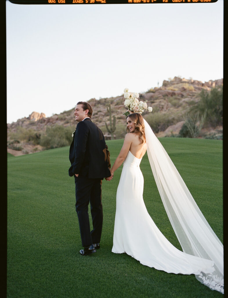 Bride and groom holding hands, smiling, in grass field in front of desert landscape.