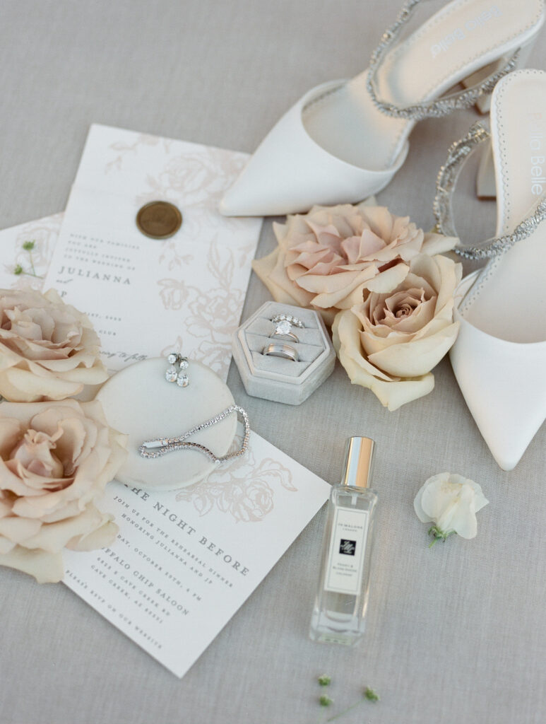 Wedding details including bride's shoes, perfume, jewelry and invitation with blush colored roses.
