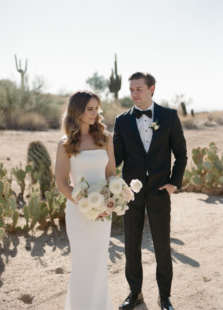 Groom looking at bride who is holding a bouquet and bride looking off to side standing in desert landscape.