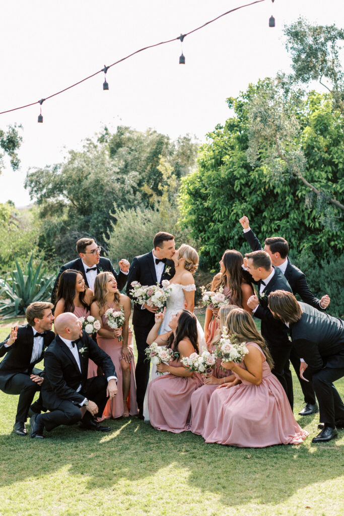 Wedding party of groomsmen in black suits and bridesmaids in blush dresses surrounding bride and groom kissing.