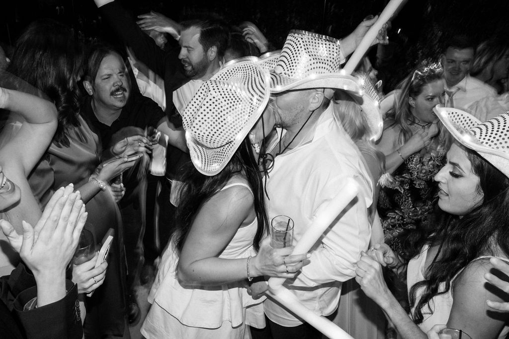 Bride and groom wearing white sequin cowboy hats, kissing at wedding reception in middle of dancing people.