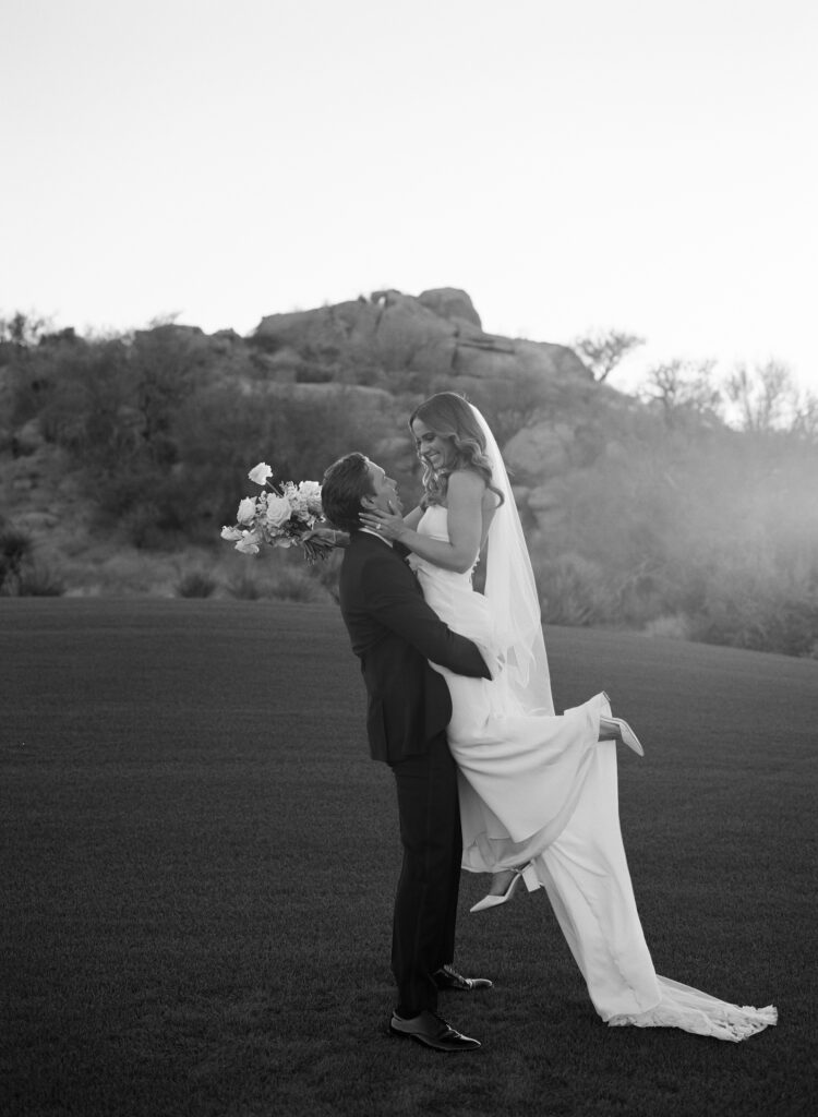 Groom lifting up bride who is holding bouquet on grass in front of desert landscape.