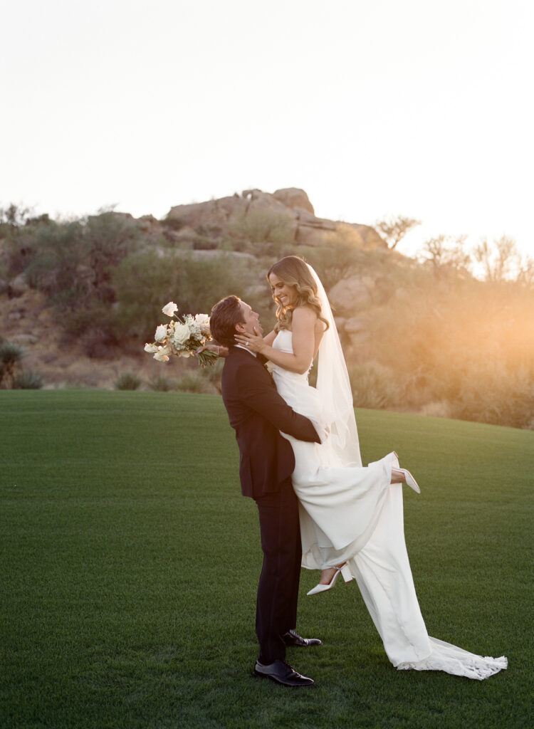 Groom lifting up bride who is holding bouquet on grass in front of desert landscape.