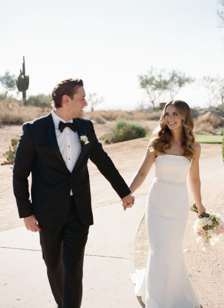 Bride and groom walking on paved sidewalk, holding hands and smiling at each other in desert landscape.