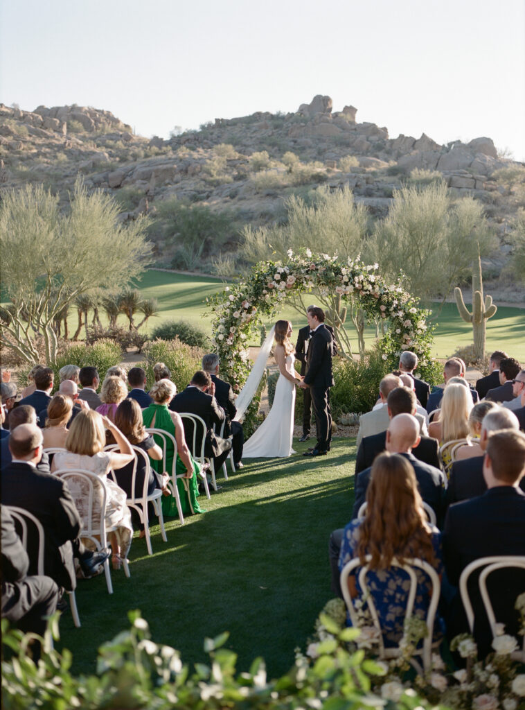 Outdoor wedding ceremony at Estancia with desert scenery and bride and groom standing in front of large floral and greenery arch behind couple.