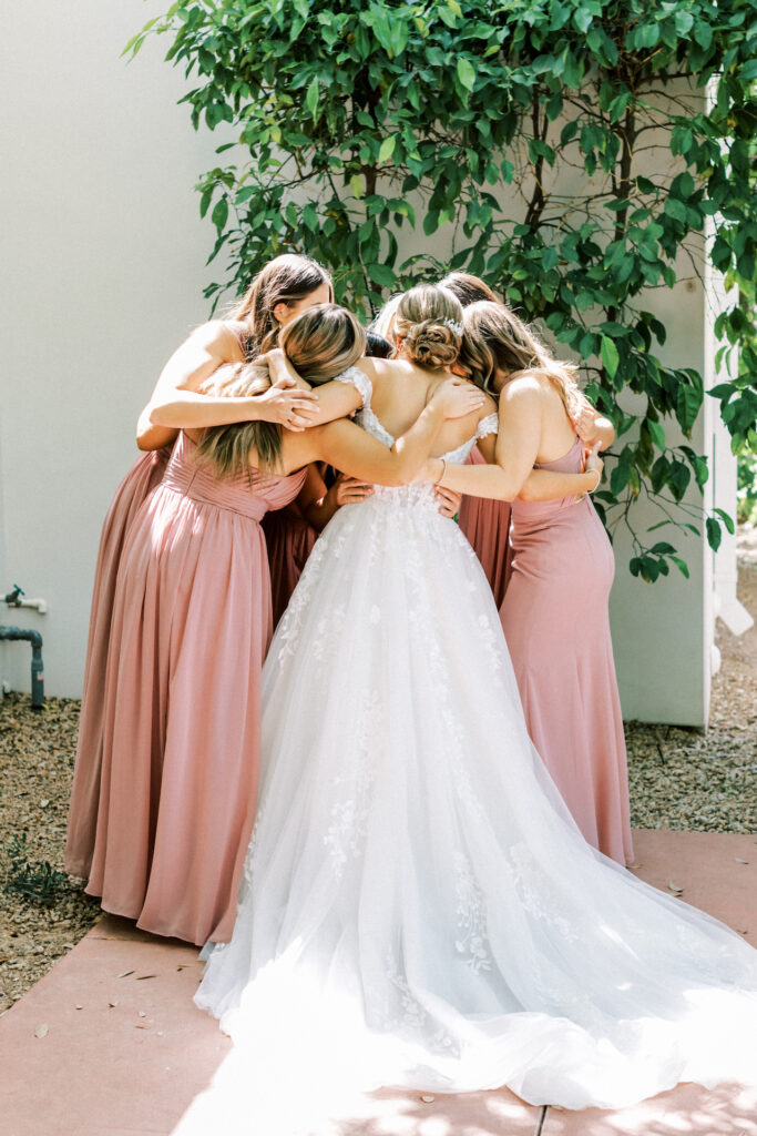 Bride with bridesmaids in blush gowns with arms around each other in a circle.