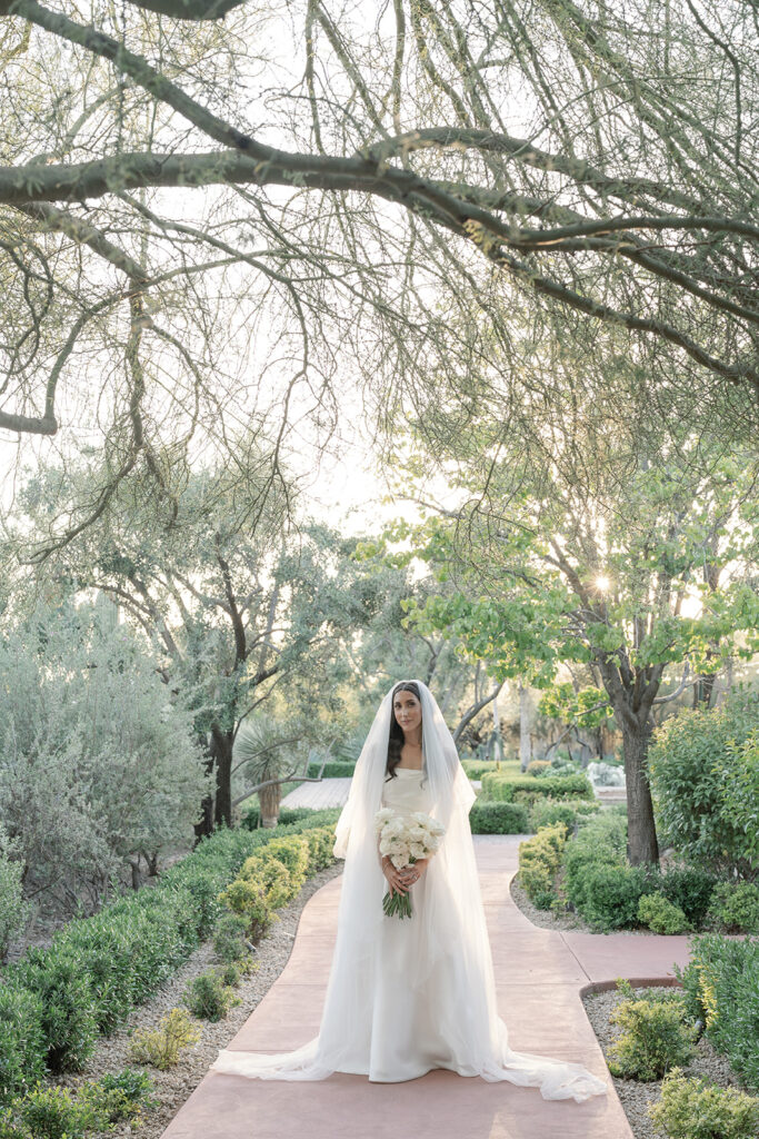 Bride standing on paved path at El Chorro, holding long stem white roses bridal bouquet.