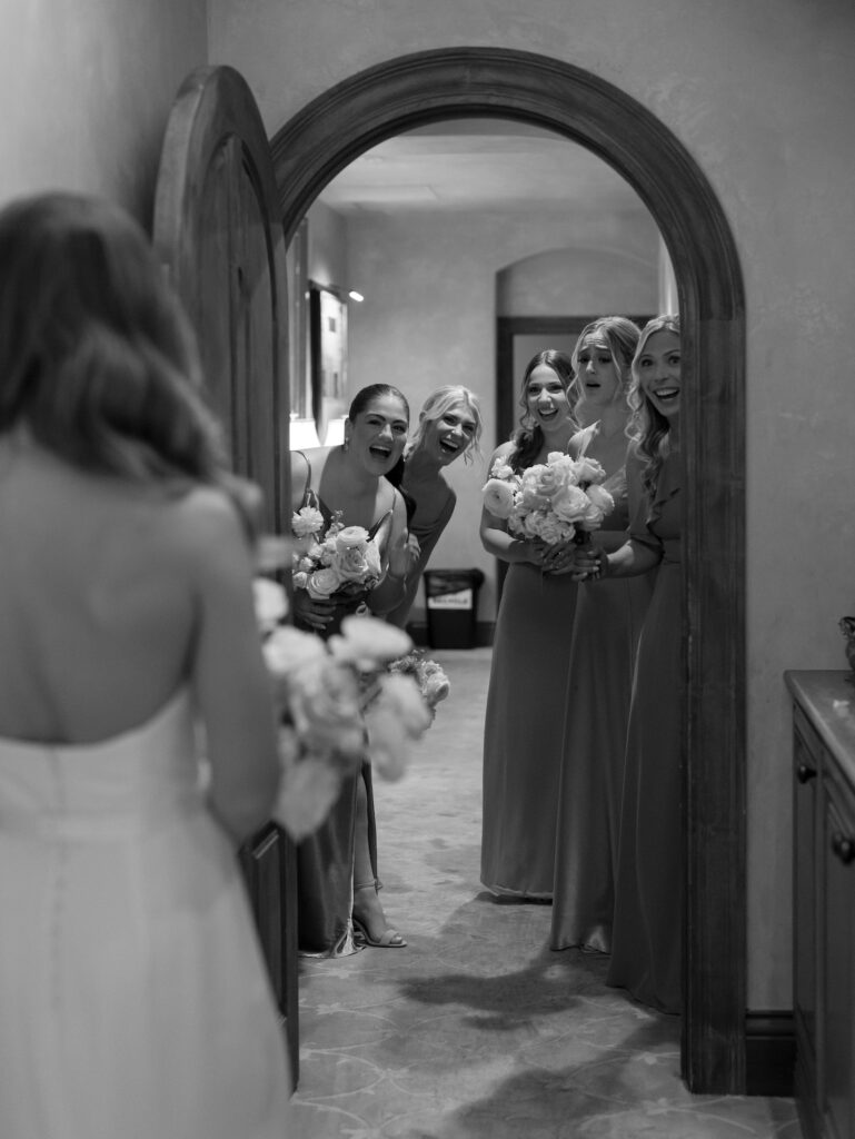 Bridesmaids looking through doorway at bride, smiling for first look in her wedding dress.