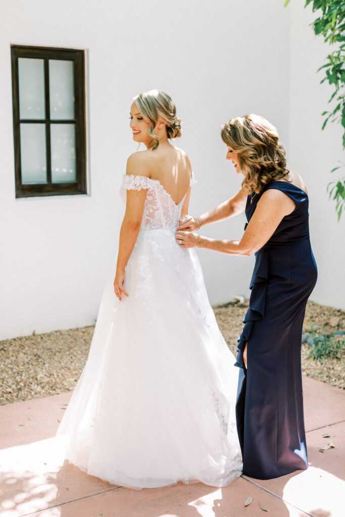 Woman buttoning up bridal gown on bride outside.