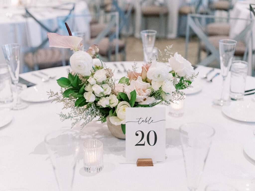 Wedding reception table with white linen and a floral centerpiece of white and blush floral with a table number sign and votive candles.