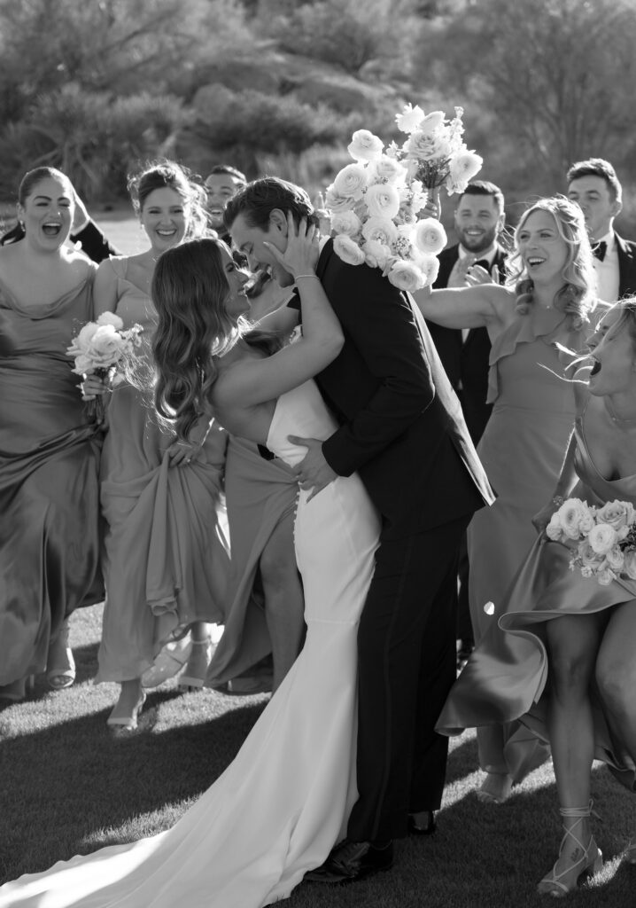 Bride and groom embracing, smiling in front of wedding party of bridesmaids and groomsmen celebrating.