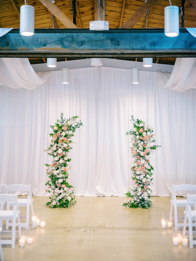 Indoor wedding ceremony floral pillars of pink and white with greenery at ceremony altar space.