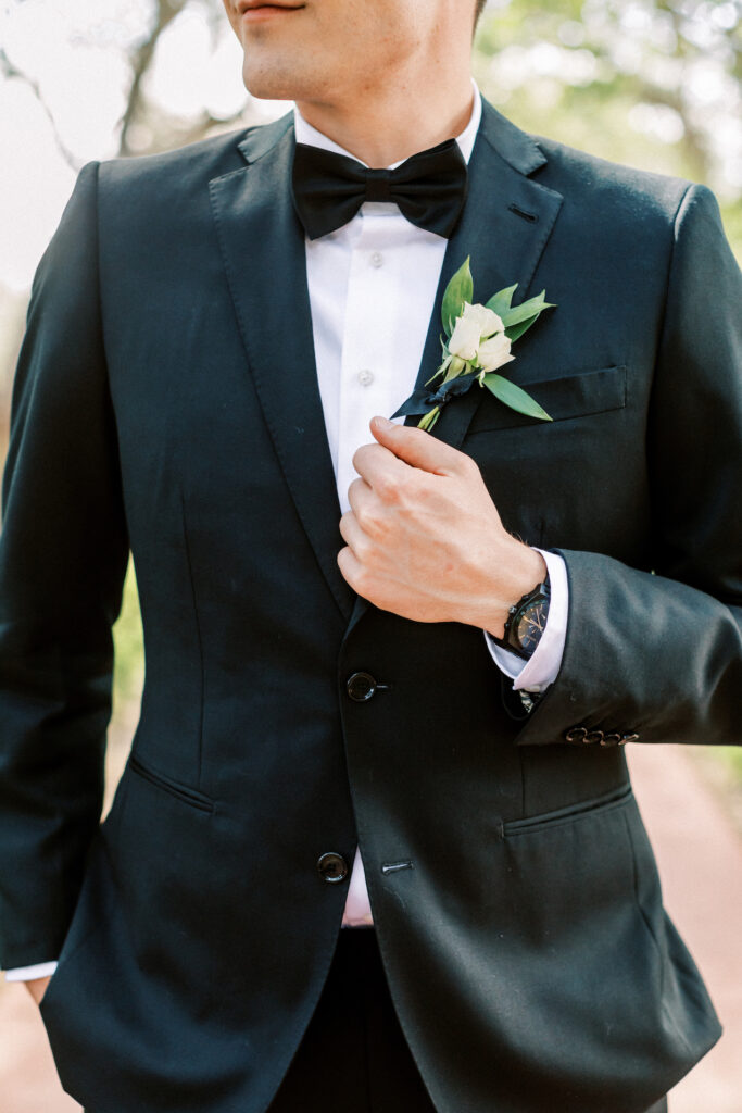 Groom boutonniere of small what flowers and greenery on his black suit with bow tie.