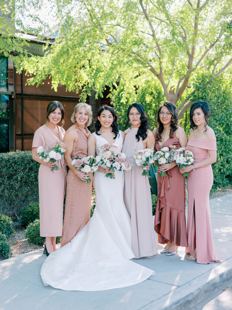 Bride standing in a line with bridesmaids outside all holding bouquets of white and blush, pink flowers and bridesmaids wearing shades of pink gowns.