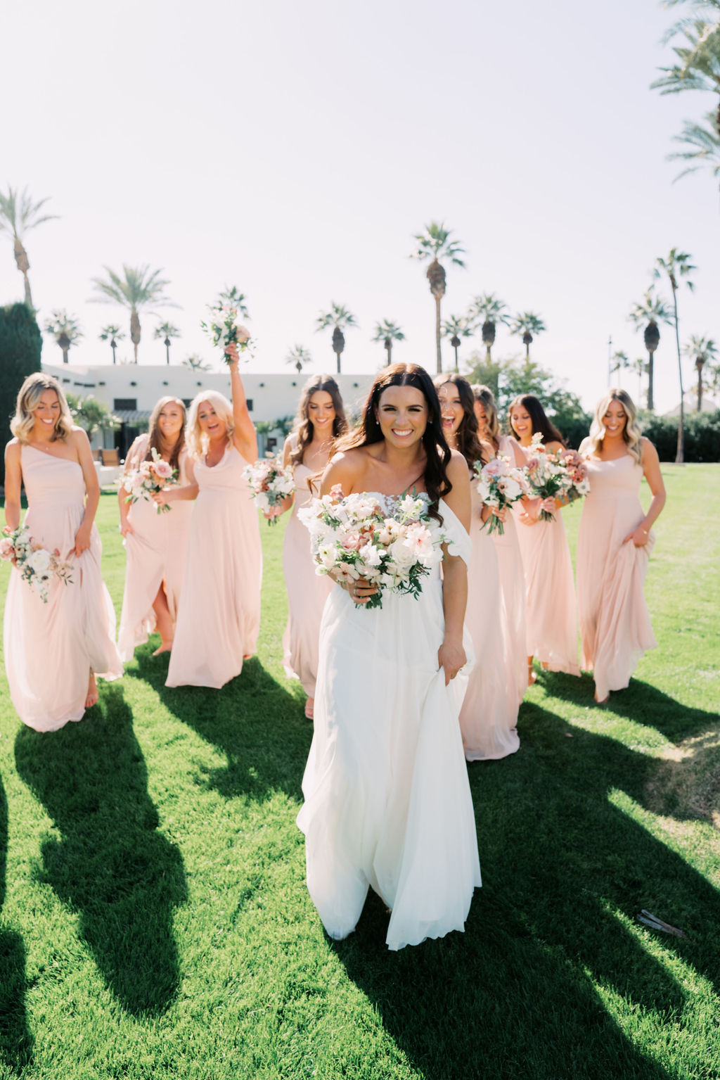 Bride walking in front of bridesmaids in blush dresses on grass field, all smiling.