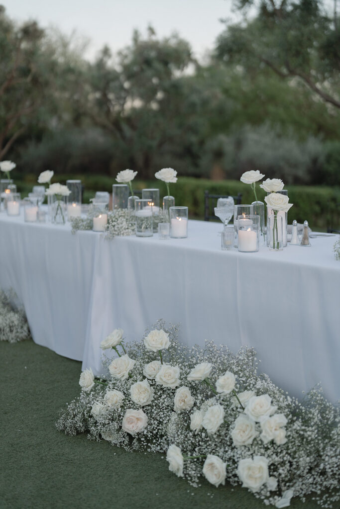 Long reception table with white linens and white roses in bud vases with baby's breath on table on on ground in front.