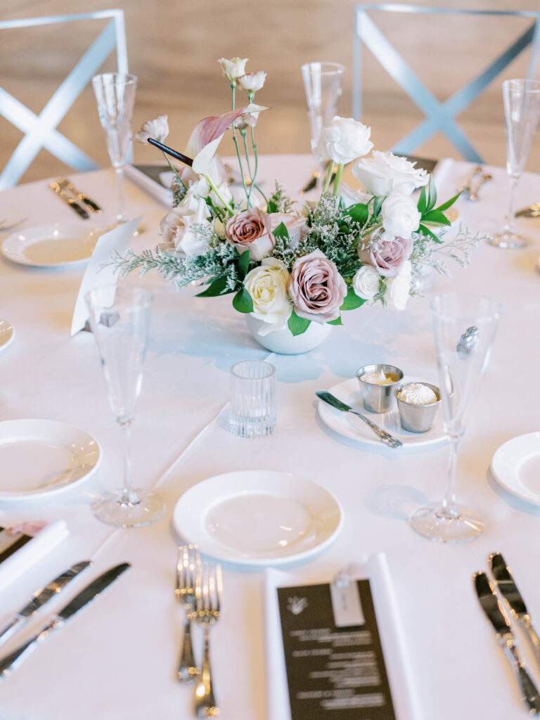 Floral centerpiece in white vase of white and blush flowers and greenery on table of white linen at wedding reception.