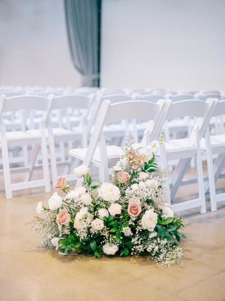 Back of aisle floral arrangement of white and blush flowers with greenery at indoor wedding ceremony.