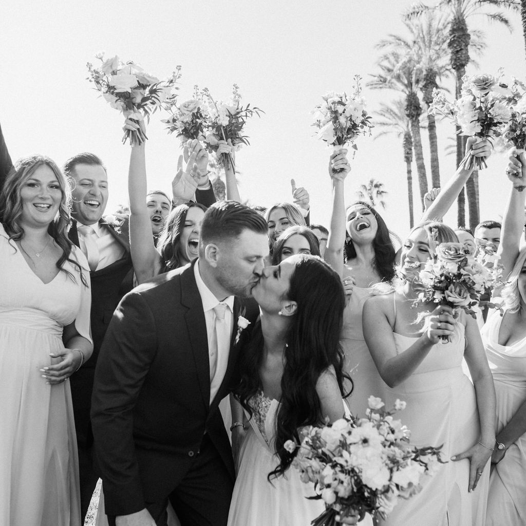 Black and white picture of wedding party outside with bride and groom at center kissing, all smiling.