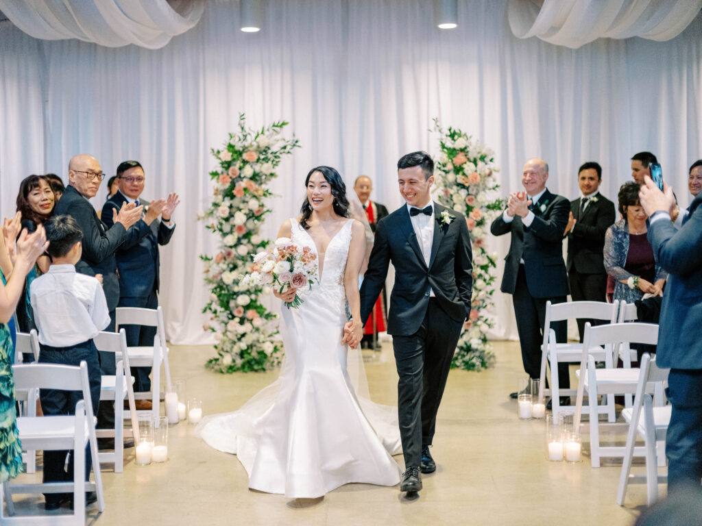 Bride and groom holding hands walking down indoor wedding ceremony aisle, smiling, after ceremony.