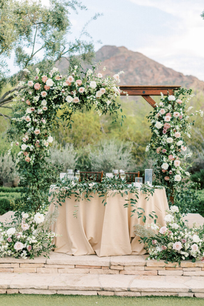 Outdoor wedding reception sweetheart table with floral installation arch behind and ground floral in front and candles with greens on table.