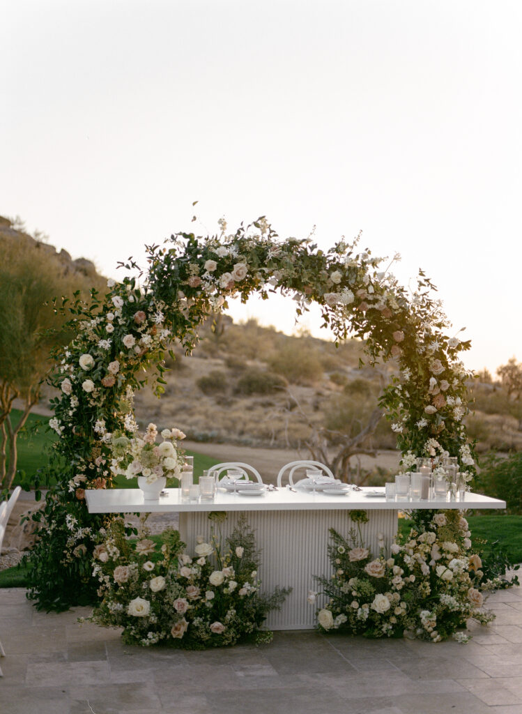 Outdoor wedding reception sweetheart table with garden arch of floral and greenery behind it and ground floral in front.