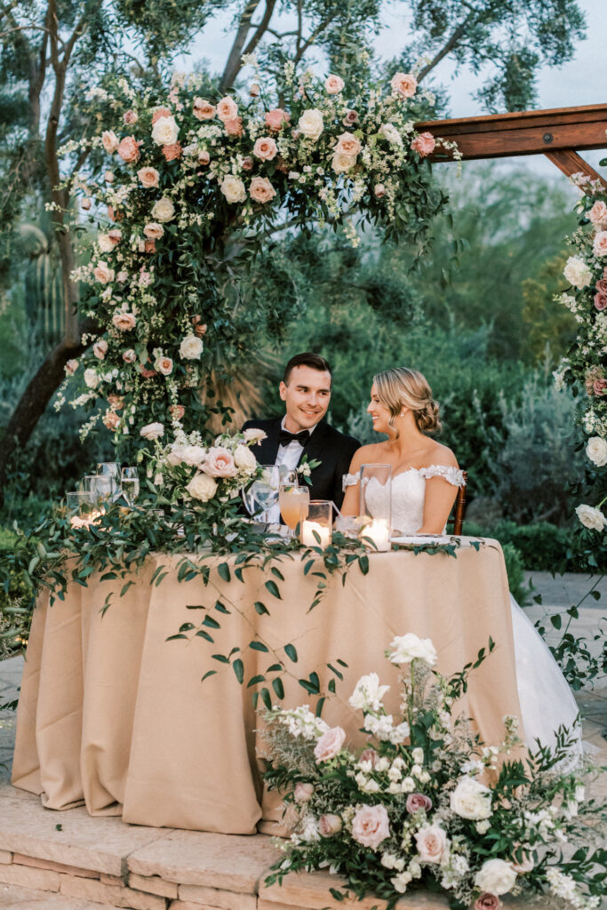 Bride and groom sitting at outdoor wedding reception sweetheart table with floral installation arch behind and ground floral in front and candles with greens on table.