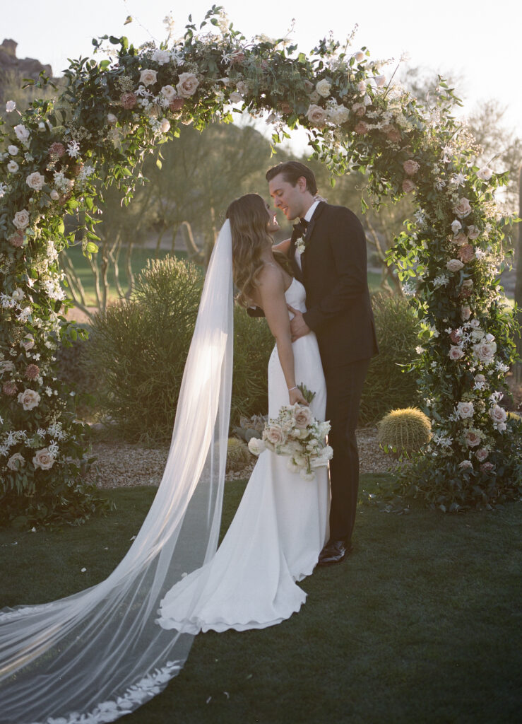 Bride and groom embracing under wedding ceremony arch of floral and greenery.