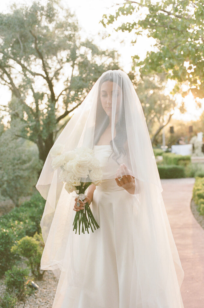Bride with veil over her face wearing wedding gown and holding long stem white roses bouquet.
